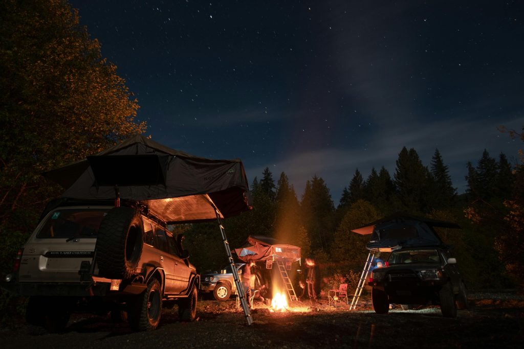 Camping scene with cars and tents under a starry night sky, featuring a cozy bonfire in the forest.
